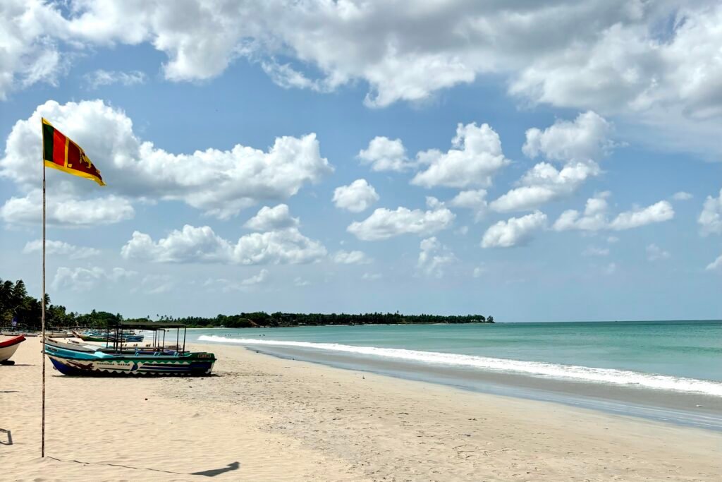 Uppuvelli Beach in Trincomalee on a sunny, slightly cloudy day. Boats are parked on the sand next to a Sri Lankan flag.