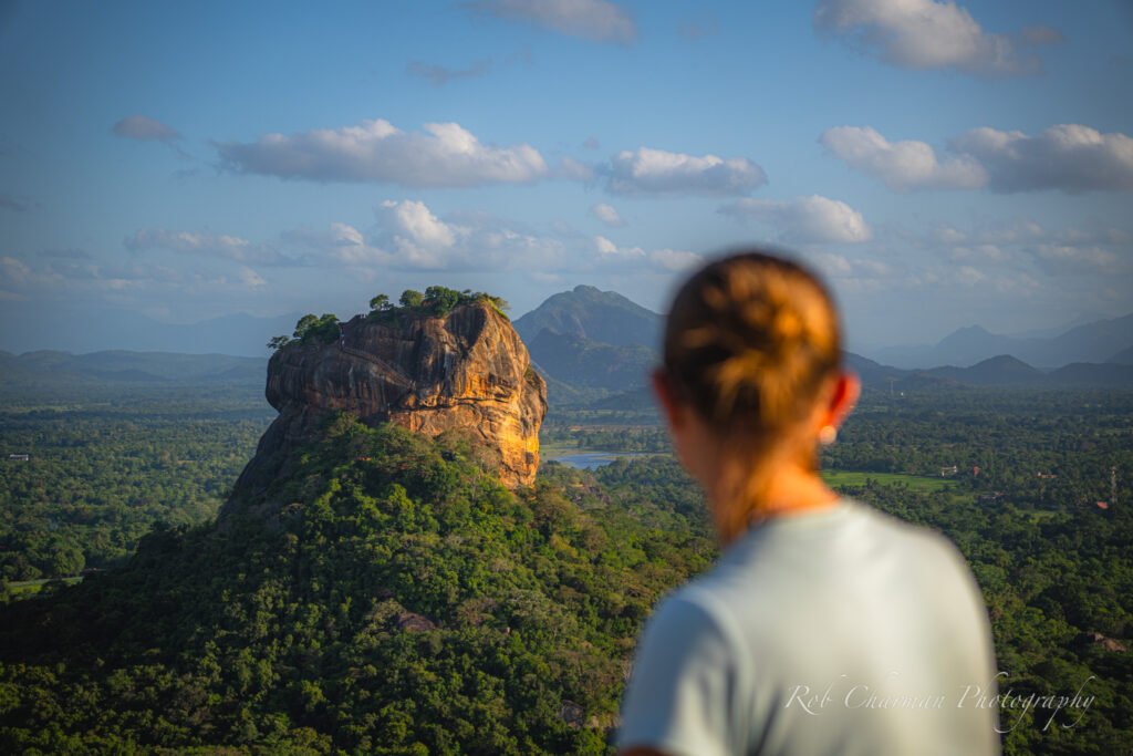 Woman overlooking Lion Rock in Sigiriya. Green forest surrounds the rock with mountains in the background. Photographed from Pidurangala Rock. 